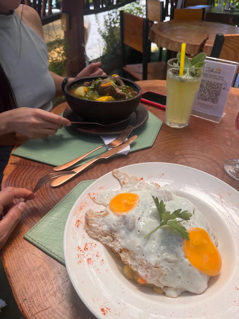 Dos personas almorzando: cazuela, huevo frito y limonada sobre la mesa de madera.