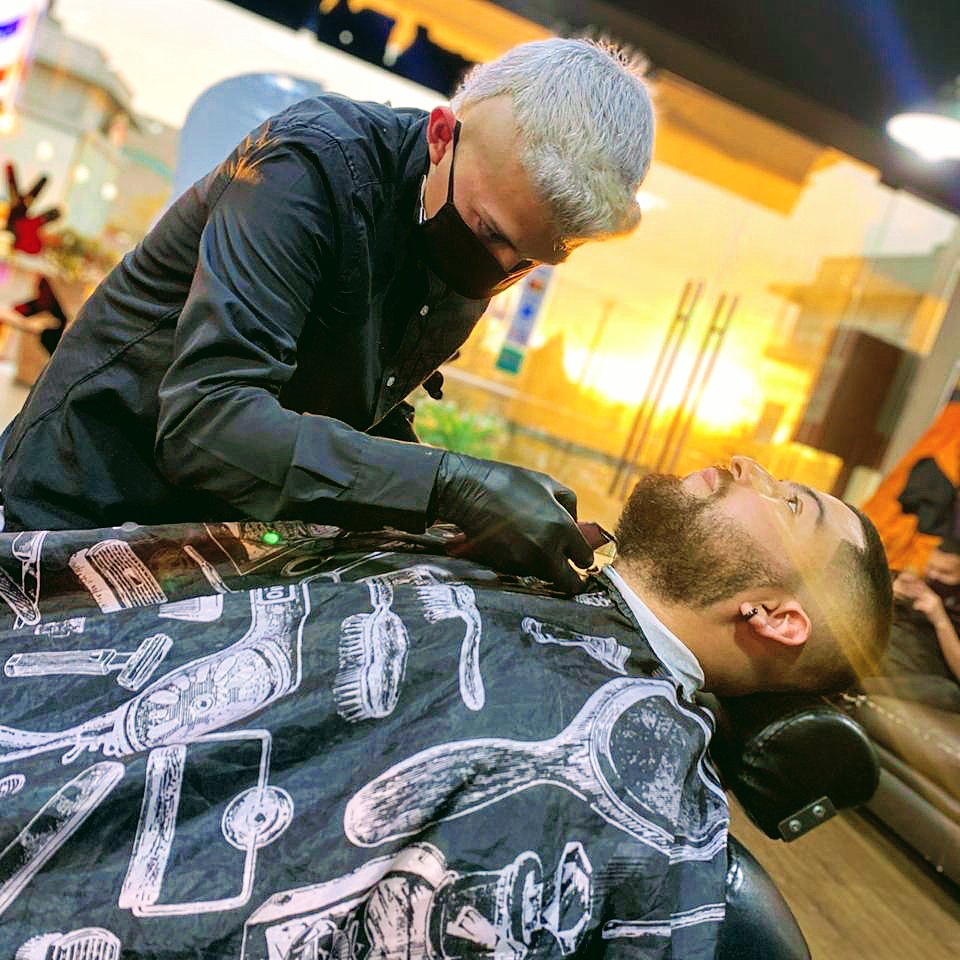 Barbero perfilando la barba de un cliente reclinado en el sillón, con luz de atardecer entrando por la vitrina