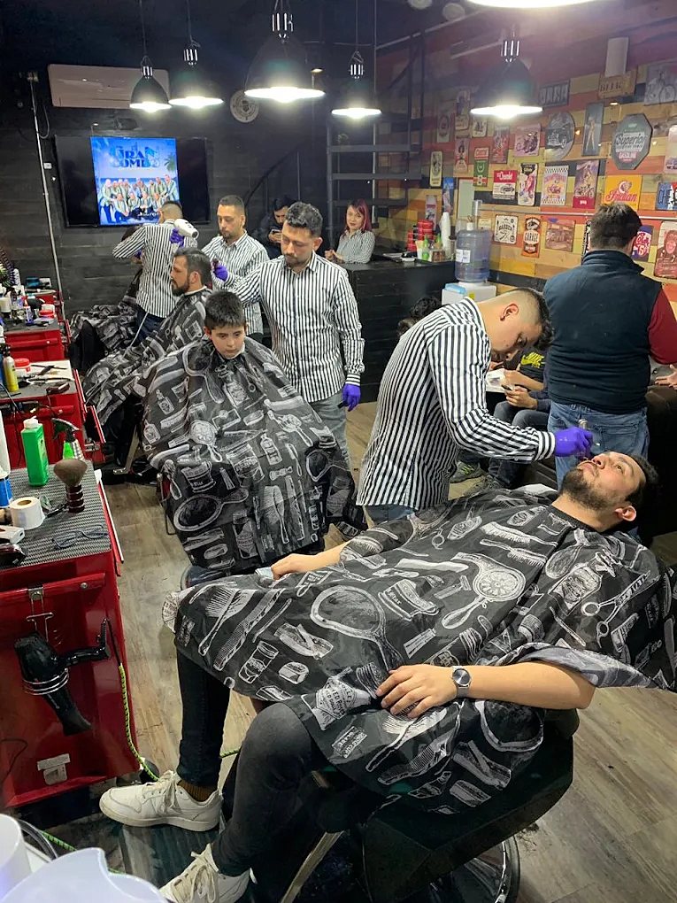 La barbería llena: varios barberos atendiendo a la vez y clientes esperando