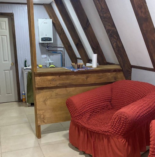 Interior de la cabaña: sillón tapizado en rojo tierra, vigas de madera y muro de la escalera.