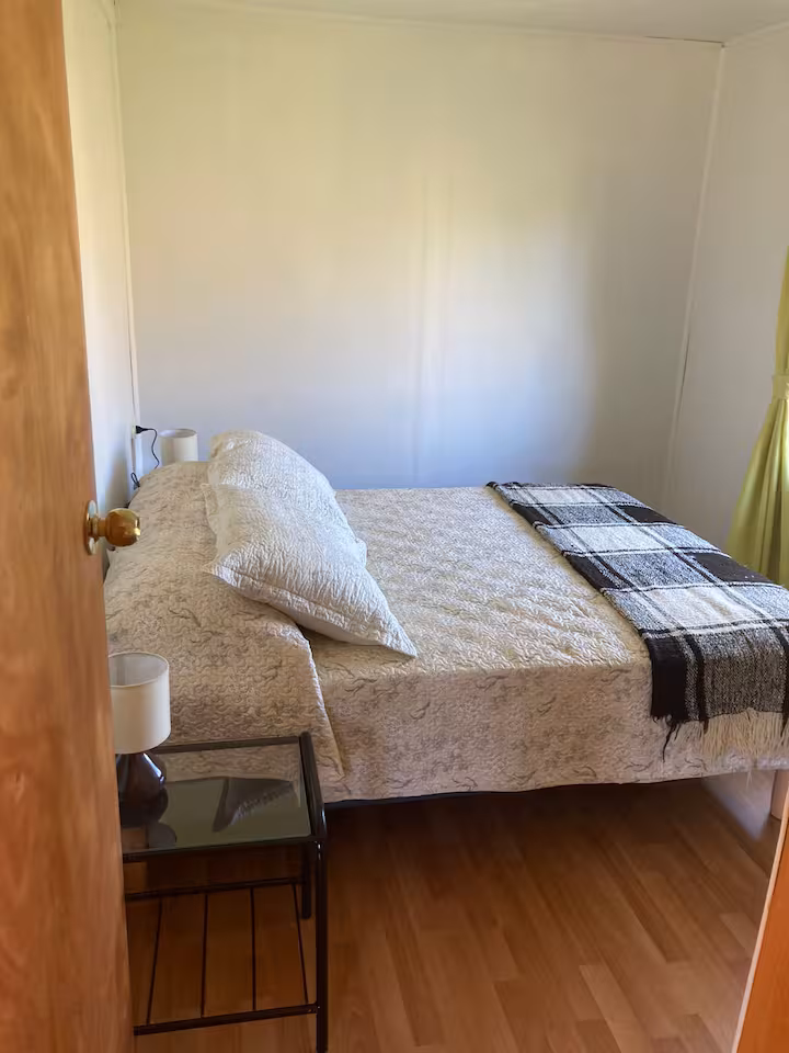 Cama tendida junto a una ventana, con velador de vidrio