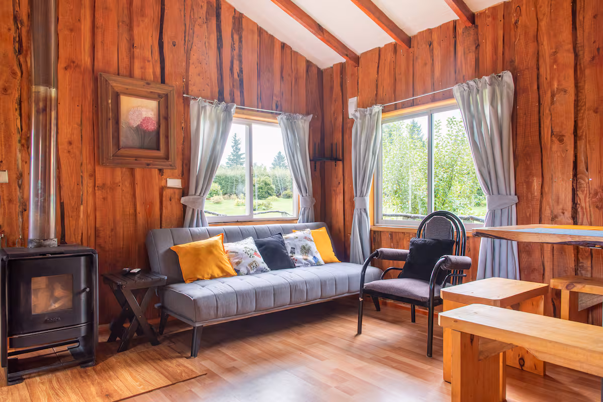 Living de la cabaña con estufa a leña, sofá gris y mesa de madera junto a la ventana