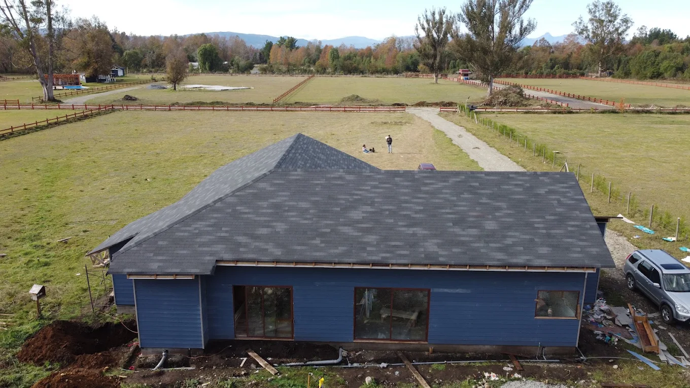 Vista aérea de una casa terminada con techo de asfalto shingle y revestimiento azul, en un predio con praderas al fondo.