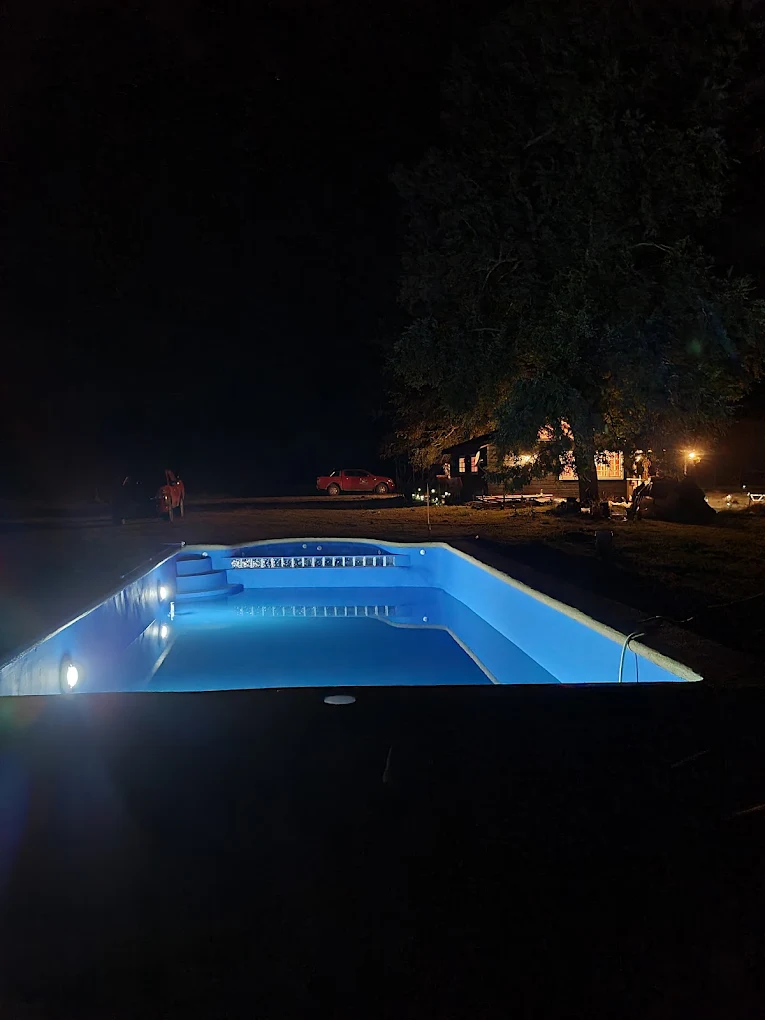 Piscina iluminada de noche vista desde lejos, con la casa encendida al costado.