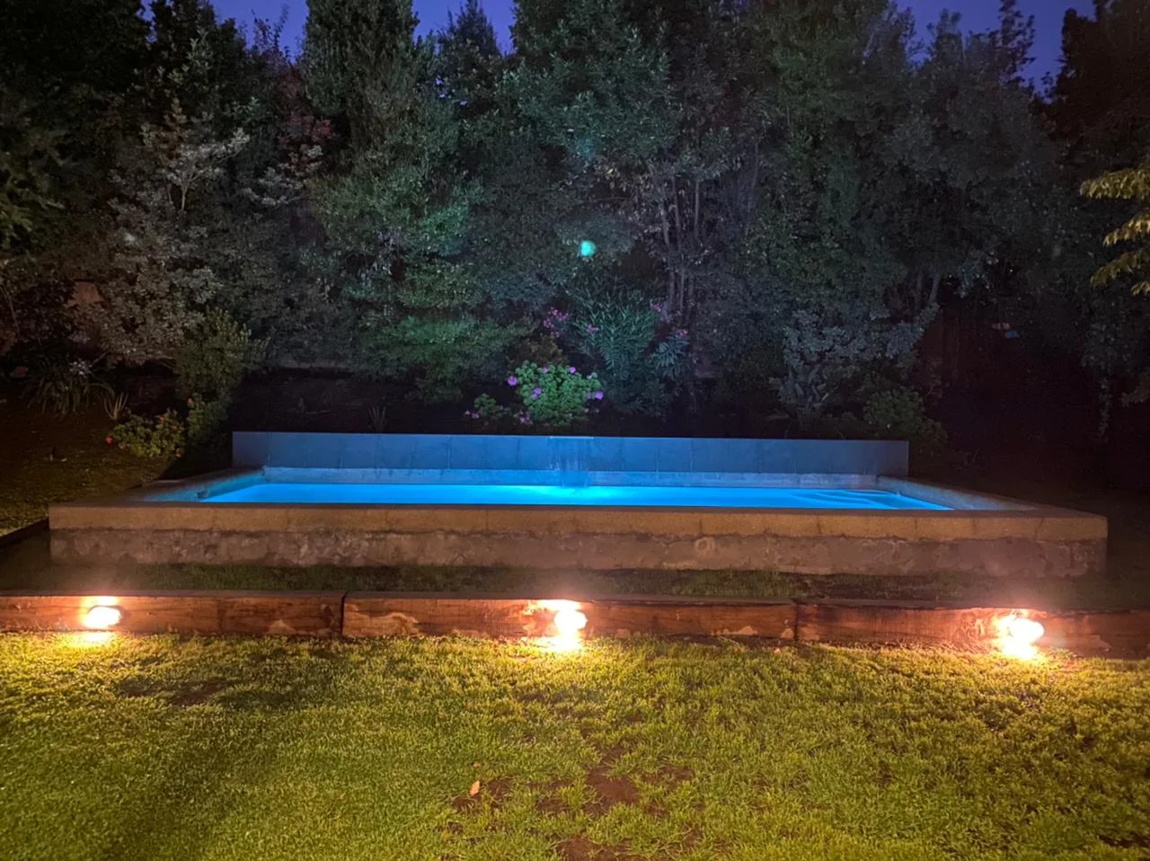 Piscina terminada e iluminada de noche, con el borde de hormigón y luces bajas en el pasto.