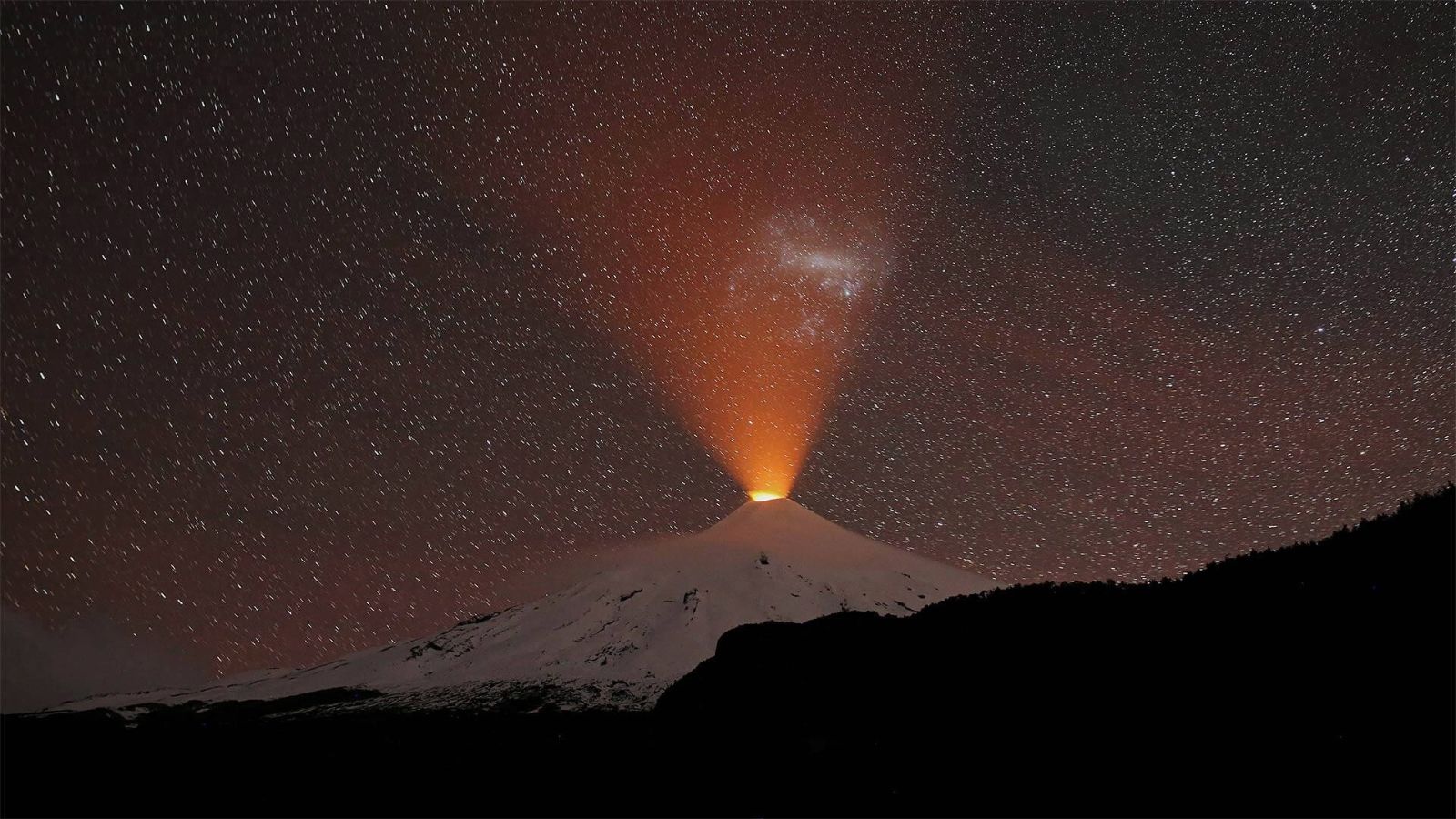 El volcán Villarrica de noche bajo un cielo estrellado, con la nieve iluminada y una columna de lava naranja saliendo del cráter.