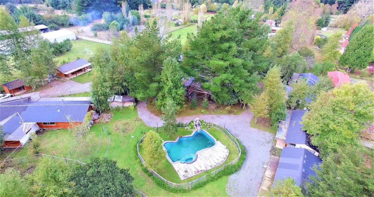 Vista aérea del centro turístico: la piscina con su cascada al centro, rodeada de cabañas de madera y árboles.
