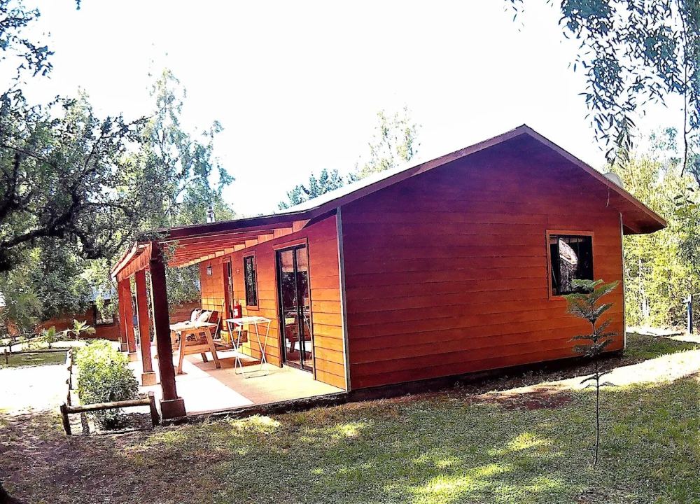 Cabaña de madera con techo de dos aguas y una terraza cubierta con mesa y sillas, rodeada de pasto y árboles.