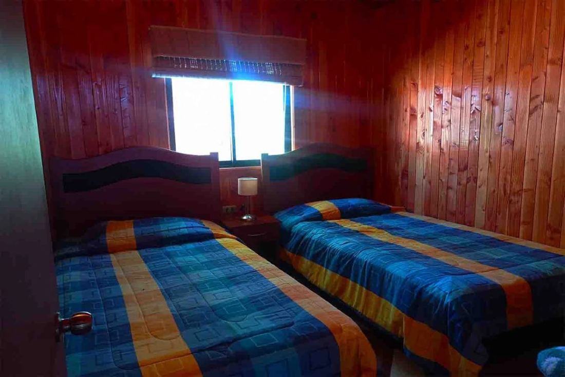 Dormitorio con dos camas de una plaza, muros de madera y una ventana entre los respaldos.