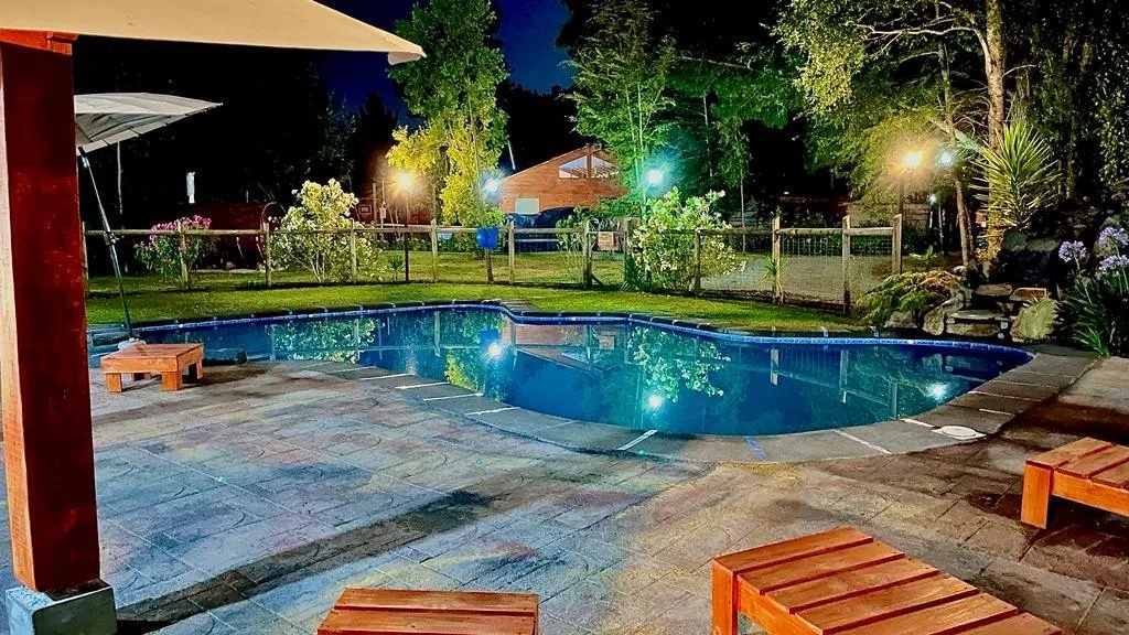 La piscina exterior iluminada de noche, con tumbonas de madera alrededor y el jardín encendido.