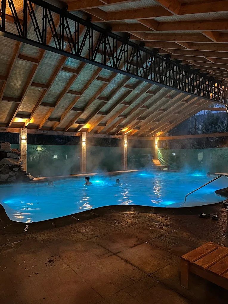 Piscina temperada bajo un techo de vigas de madera, con vapor sobre el agua iluminada de azul y gente bañándose de noche.