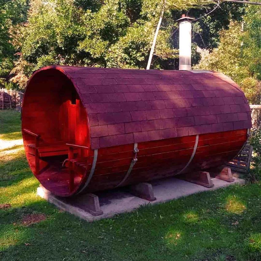 Sauna con forma de barril de madera y chimenea metálica, sobre el pasto entre los árboles.