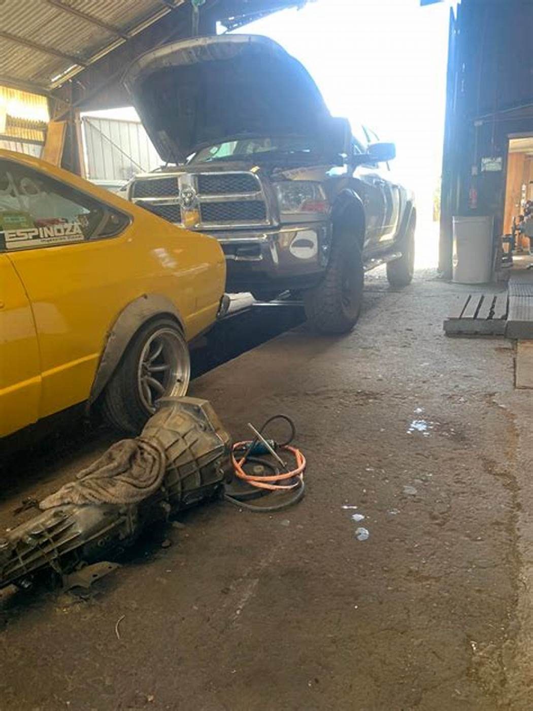 Interior del taller con varios autos y una caja de cambios desmontada en el suelo.