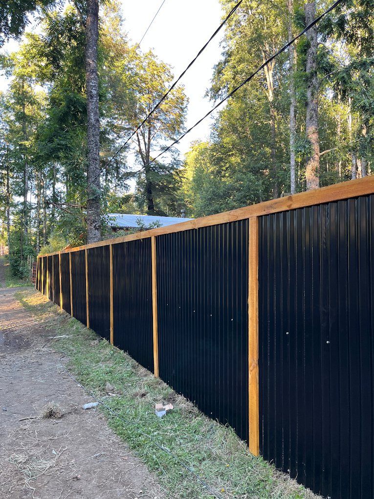 Cerco perimetral largo de tablas oscuras con pilares de madera a la vista, junto a un camino.