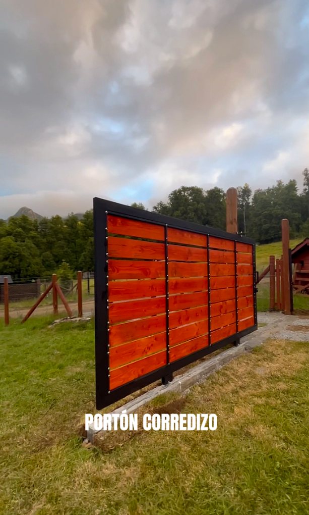 Portón corredizo de tablas de madera barnizada con marco de fierro negro, instalado en la entrada de una parcela.